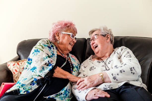 Senior Friends Having a Good Laugh Two senior woman sitting chatting in a nursing home in North East of England. One has pink hair. They are sitting on a sofa having a good laugh. One woman is wearing a nasal oxygen cannula. Another lady has a personal alarm GPS tracker around her neck DisruptAgingCollection stock pictures, royalty-free photos & images
