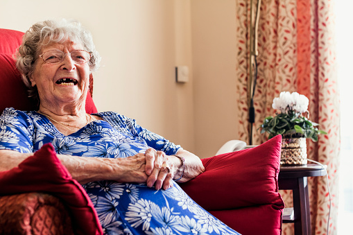 Senior woman laughing while sitting in an armchair in a nursing home in North East of England. She has her walking aid in front of her.