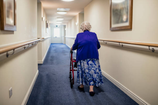 Senior Woman with Walker in a Care Home A senior woman walking down a corridor with the assistance of a walker. view from rear assisted living stock pictures, royalty-free photos & images