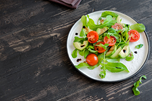 Avocado and tomatoes salad with arugula and balsamic vinegar over wooden, copy space. Healthy diet vegan vegetarian summer vegetable salad food concept.