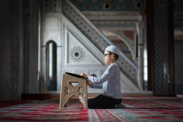 A young Muslim boy wearing a blue T-shirt and black Taqiyah cap poses for a photo reading a book in a park. Soft and selective focus.