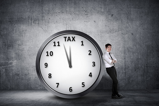 Happy asian businessman lean back on the clock with deadline time for paying tax. Goods and Services Tax concept.