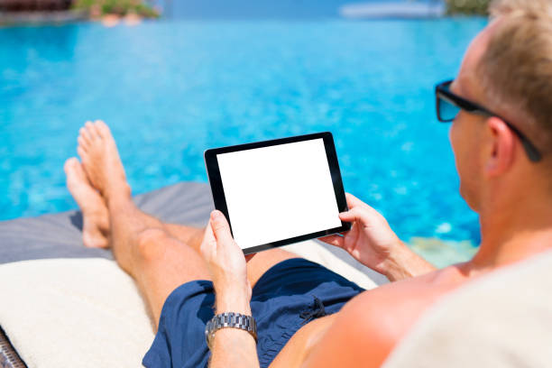 istock Man using tablet by the pool, blank white mock up screen in horizontal orientation 938505032