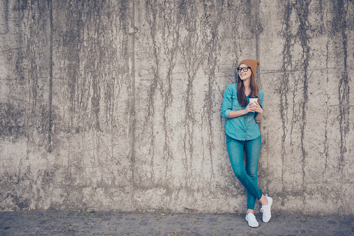 Full size of cheerful young lady, standing near concrete wall outside, smiling, with crossed legs and plastic cup with coffee, in casual jeans outfit and brown hat, glasses