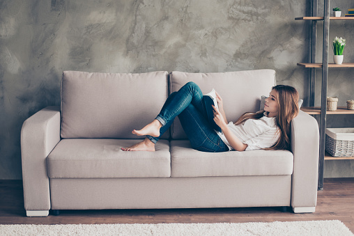 Domestic life. Relaxed young lady is studying, lying on the cozy beige couch in living room at home, so nice modern interior, so comfortable atmosphere for study and work