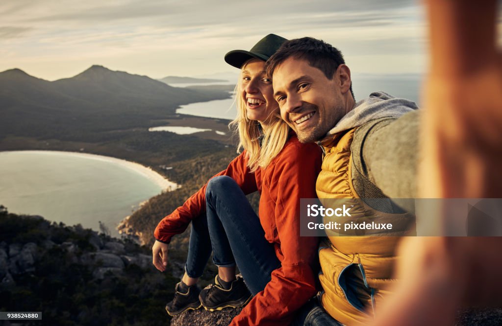 The perfect backdrop for our love Cropped portrait of an affectionate young couple taking selfies while sitting on a mountain peak Couple - Relationship Stock Photo