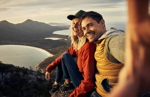 The perfect backdrop for our love Cropped portrait of an affectionate young couple taking selfies while sitting on a mountain peak backpack photos stock pictures, royalty-free photos & images
