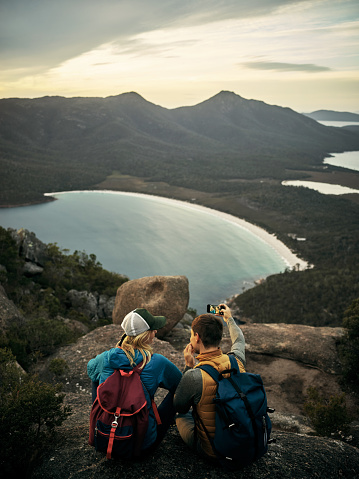 Rearview shot of a young couple taking selfies while sitting on a mountain peak