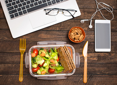 Healthy business lunch at workplace. Salad, salmon, avocado and nuts lunch box on working desk with laptop, smartphone, glasses and headphones.