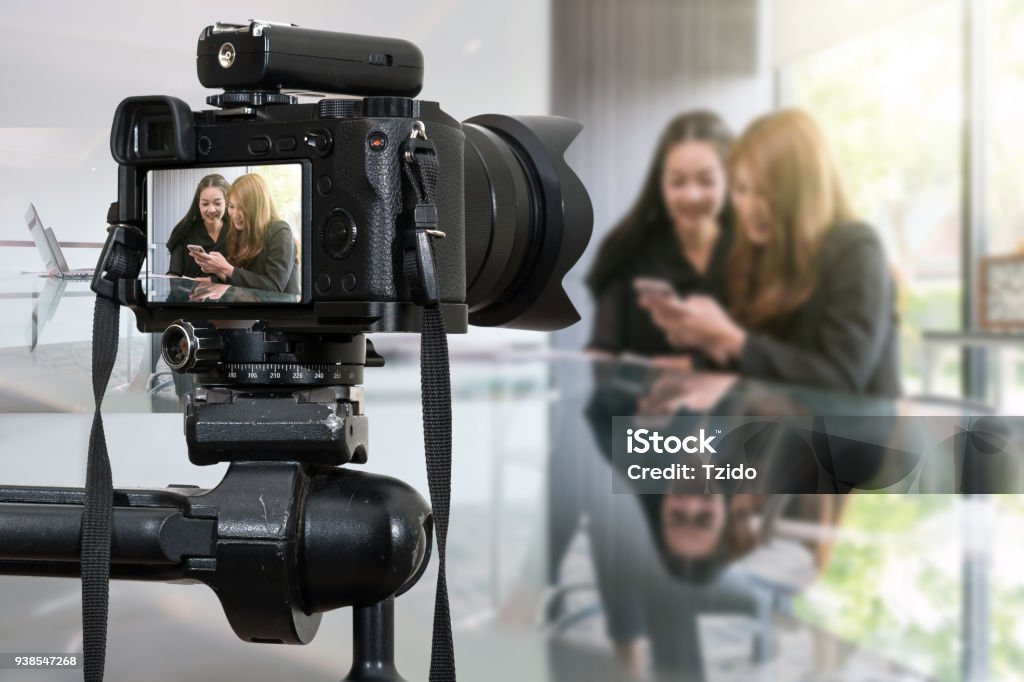 Cámara sin espejo digital profesional con micrófono de grabación de vídeo blog de dos asiáticas joven mujer de negocios usando el teléfono inteligente, la cámara para el fotógrafo o el concepto de vídeo y transmisión en vivo - Foto de stock de Cámara cinematográfica libre de derechos