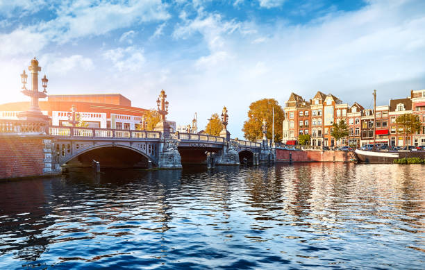 Panorama of Blue Bridge (Blauwbrug) in Amsterdam on river Amstel. View to building of National opera and ballet at sunset. Blue sky with clouds, street lamps on bridge. Evening landscape.