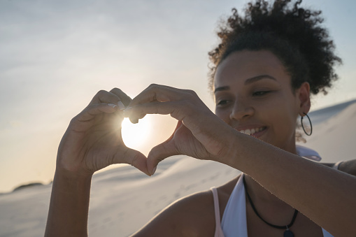 Portrait of a loving woman at the desert making a heart shape with the sunset at the background and looking happy