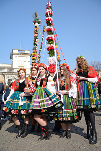Łowicz, Poland - March 25th, 2018: Team of teenage girls in folk costumes on Łowicz main square during a competition for the most beautiful Easter palm. Some big palms behind the girls, over blue sky. Town hall in the background. Vertical image in a sunny day.