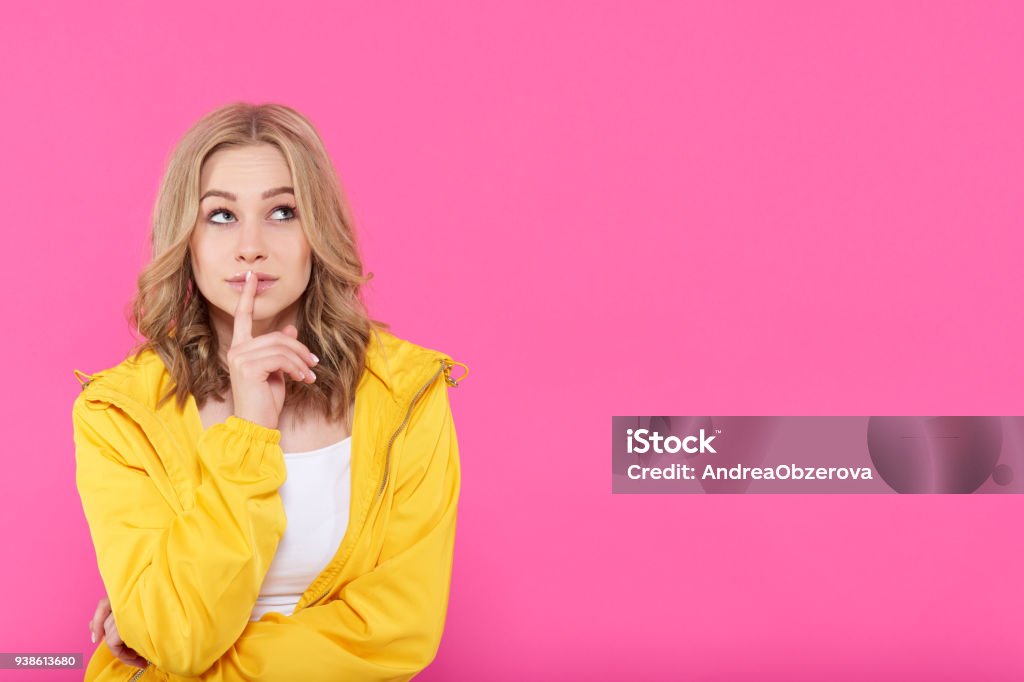 Beautiful trendy girl in colorful clothes deep in thoughts, looking up with finger on lips. Attractive young woman portrait over pastel pink background. Imagination concept. One Woman Only Stock Photo