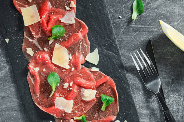 Top view of the raw beef carpaccio with parmesan cheese on a black shale service board. Delicatessen food. Top view of the raw beef carpaccio with parmesan cheese on a black shale service board. Delicatessen food. carpaccio parmesan cheese beef raw stock pictures, royalty-free photos & images
