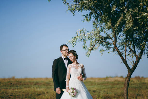 beau mariage photosession. beau marié dans un costume noir et une jeune mariée en robe de dentelle blanche avec coiffure exquis sur la promenade dans le grand champ vert dans le ciel et le grand arbre - veil bride lace married photos et images de collection