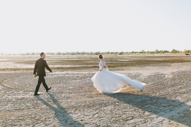 beau mariage photosession. beau marié unshaved dans un pantalon noir avec bouquet et jeune mariée mignonne en robe motif dentelle blanche avec coiffure exquis sur la promenade le long du coucher de soleil côte - veil bride lace married photos et images de collection