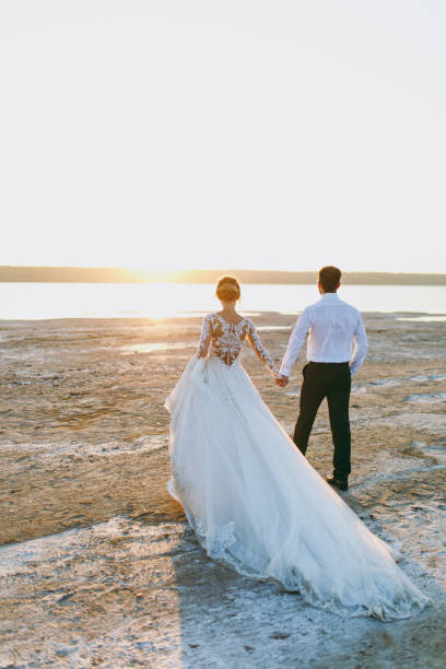 beau mariage photosession. beau marié unshaved dans un noir pantalon et la jeune mariée mignonne en point ajouré blanc robe avec coiffure exquis sur la promenade le long de la côte près du coucher du soleil de la mer - veil bride lace married photos et images de collection