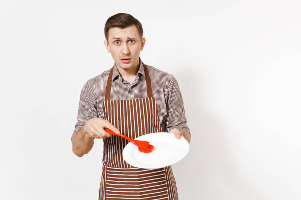 istock Young man chef in striped brown apron, shirt holds washes white round empty clear plate with red brush for washing dishes isolated on white background. Male housekeeper, houseworker or domestic worker 938641208