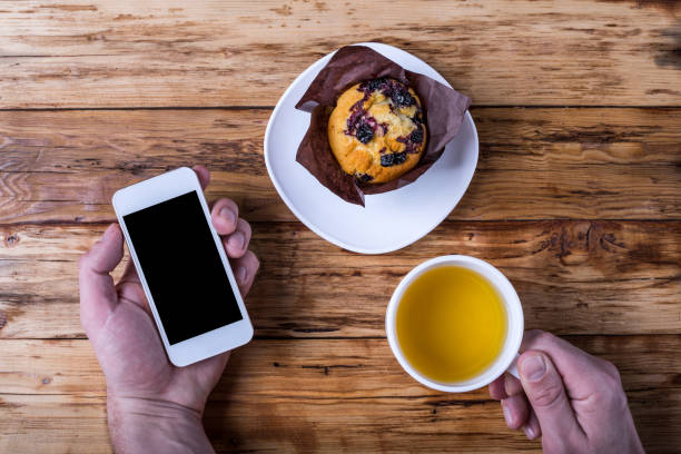 il concetto di colazione: un uomo tiene in mano uno smartphone e una tazza di tè caldo, vista dall'alto. - muffin blueberry muffin blueberry food foto e immagini stock