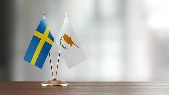 Swedish and Cypriot flag pair on desk over defocused background. Horizontal composition with copy space and selective focus.