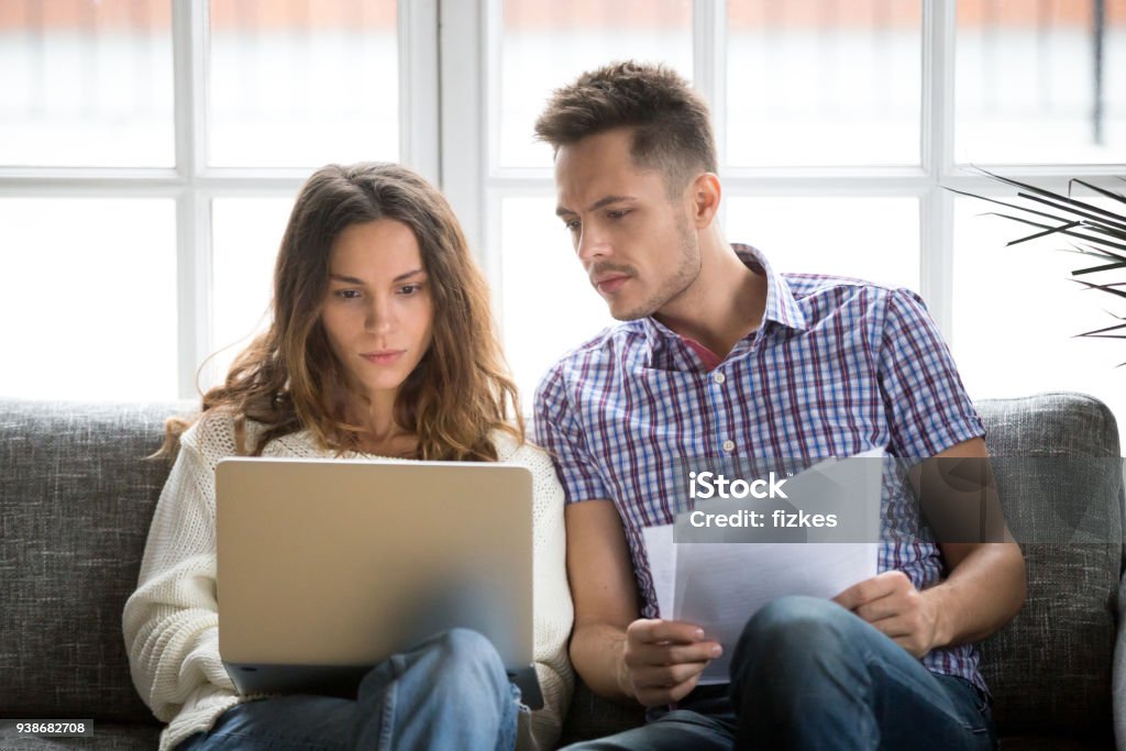 Focused worried couple paying bills online on laptop with documents Focused worried couple paying bills online on laptop with documents sitting together on sofa at home, serious confused man and woman planning budget expenses, young family having debt loan problems Couple - Relationship Stock Photo