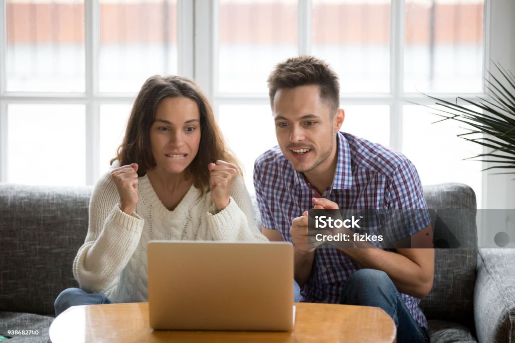 Couple looking at laptop screen supporting team watching match online Nervous excited couple in tension clenching fists looking at laptop screen, man and woman football fans cheering supporting sport team watching championship match online together hope for win at home Fan - Enthusiast Stock Photo