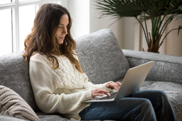 uśmiechnięta młoda kobieta siedząca na kanapie za pomocą laptopa komunikującego się online - wireless technology serious education enjoyment zdjęcia i obrazy z banku zdjęć