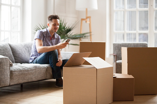 Packed cardboard boxes with young man sitting on sofa in living room calling delivery service at background, belongings in carton containers waiting for moving in out new home or relocation concept