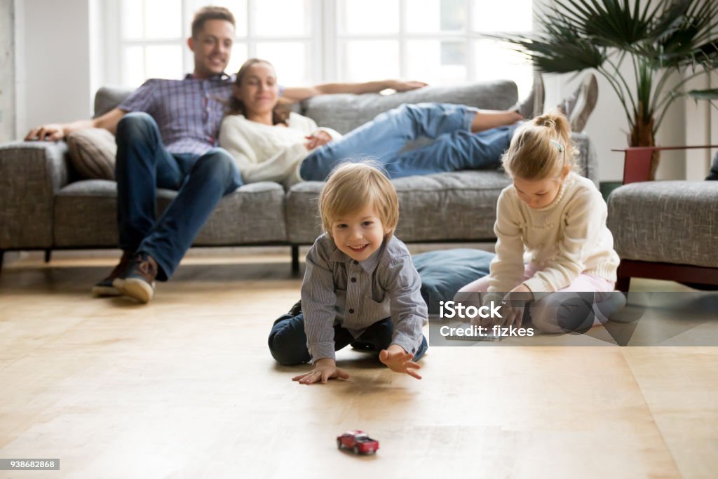 Kids playing on floor, parents relaxing on sofa at home Cute kids playing while parents relaxing sofa at home together, smiling active boy entertaining with toy car near his sister on floor, happy family spending time together in living room on weekend Flooring Stock Photo