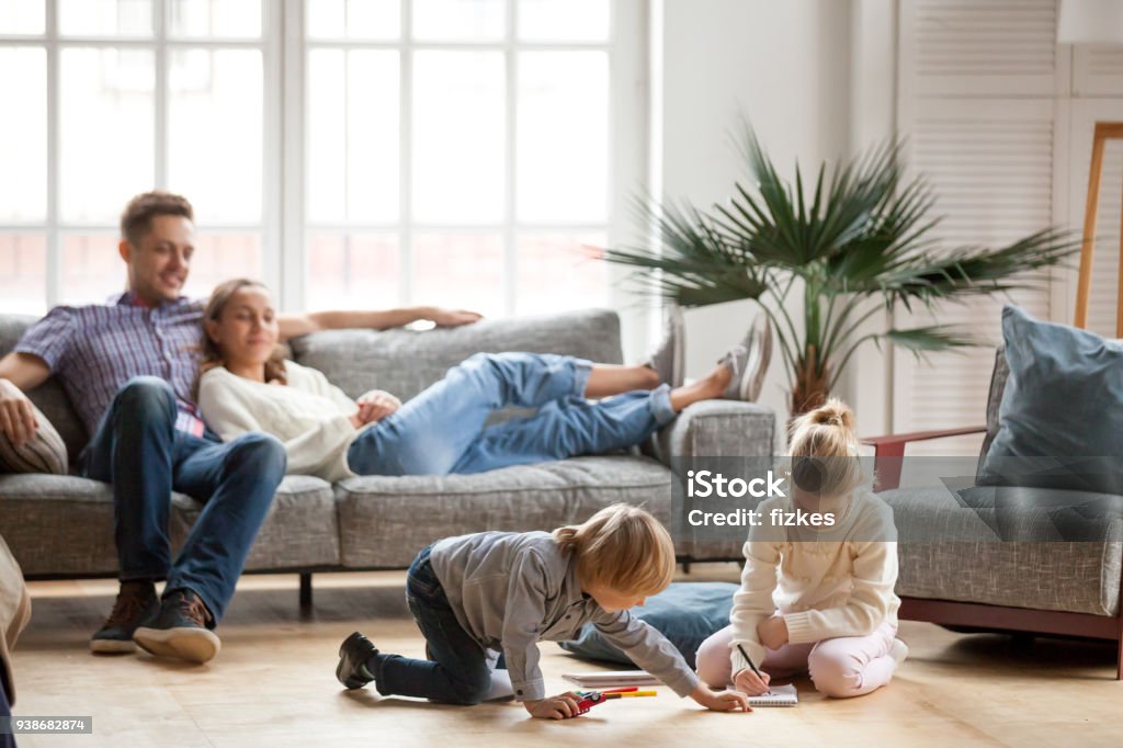 Children siblings playing drawing together while parents relaxing at home Children sister and brother playing drawing together on floor while young parents relaxing at home on sofa, little boy girl having fun, friendship between siblings, family leisure time in living room Family Stock Photo