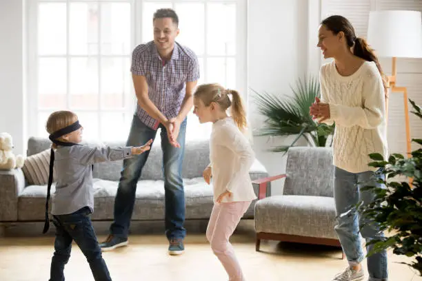 Blindfolded cute little boy playing hide and seek at home, parents and kids laughing spending time together enjoying game on weekend, happy family of four having fun leisure activity in living room