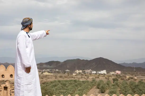 omani man in traditional outfit pointing to a distance at a countryside of Oman