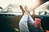Woman is holding cup of coffee inside of car. Travel lifestyle. Legs on dashboard