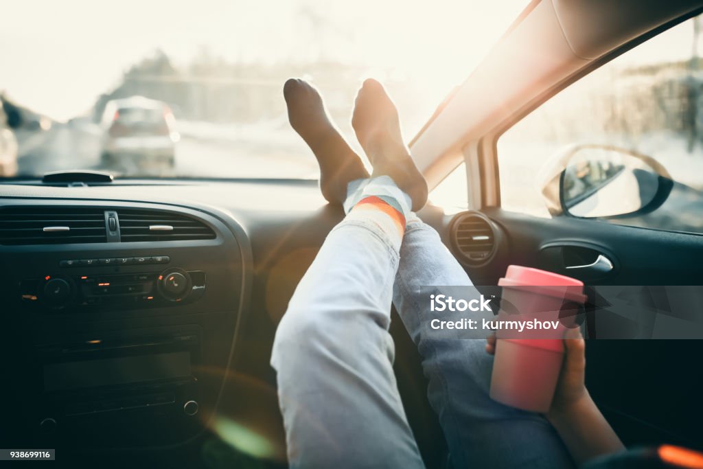 Woman is holding cup of coffee inside of car. Travel lifestyle. Legs on dashboard Car Stock Photo