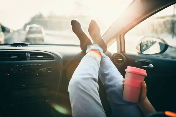 Photo of Woman is holding cup of coffee inside of car. Travel lifestyle. Legs on dashboard