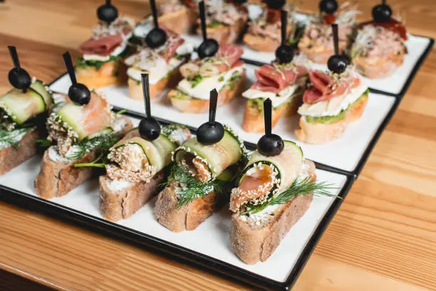 many tasty pinchos with tuna, chorizos, salmon, egg, dried tomatoes, avocado, salmon, bacon, Hamon, Brie Cheese, Olives and Pesto Sauce, traditional spanish snack, served on a white plate in a row