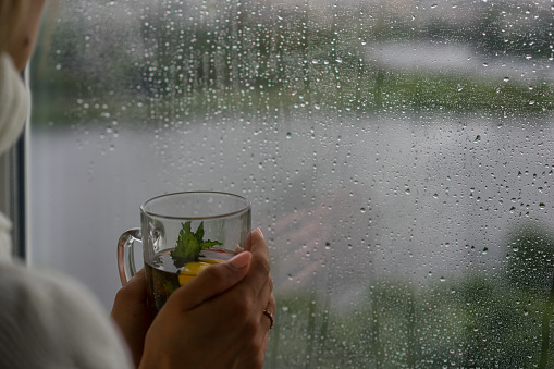 Young Woman Enjoying her morning tea, Looking Out the Rainy Window. Beautiful romantic unrecognizable girl drinking hot beverage at cozy home. Rainy Day Mood