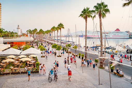 People walk on the restaurant and palm tree lined Paseo del Mullene Uno waterfront promenade in downtown Malaga Spain at sunset time, with a docked cruise ship in the background.