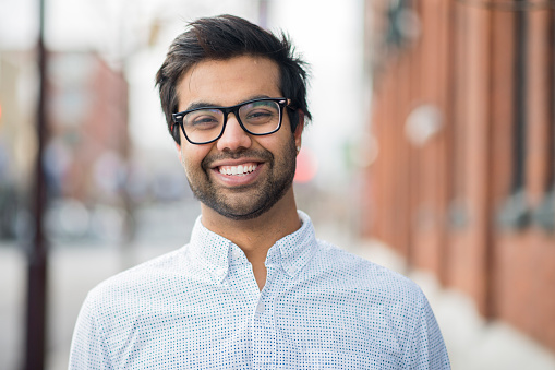An attractive young Indian man stands on the corner of a downtown city street. He smiles widely at the camera.