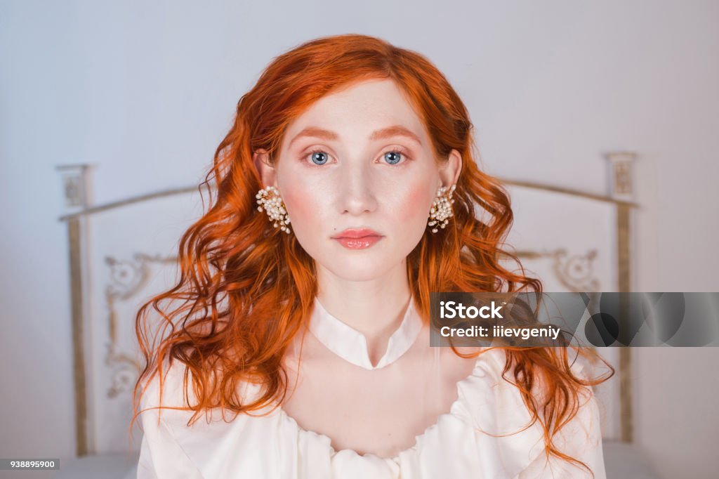 Portrait of a woman with long red curly hair in a white vintage wedding dress with white pearl earrings on her ears. Red-haired girl with a pale skin, blue eyes, a bright unusual appearance in bedroom Adult Stock Photo