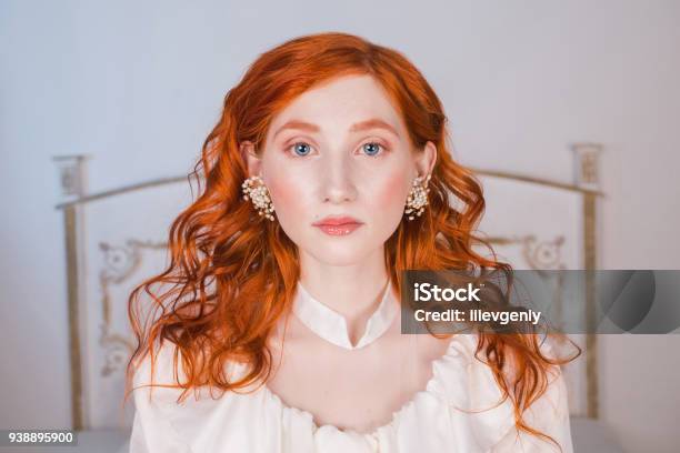 Portrait Of A Woman With Long Red Curly Hair In A White Vintage Wedding Dress With White Pearl Earrings On Her Ears Redhaired Girl With A Pale Skin Blue Eyes A Bright Unusual Appearance In Bedroom Stock Photo - Download Image Now