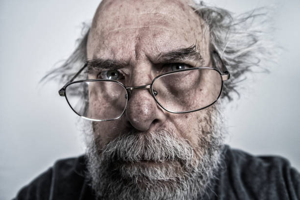 Close-up of the ominous face of a bizarre, spooky senior adult man with crazy tangled hair and a dead serious facial expression. His eyeglasses are halfway down his nose as he glares at the camera.