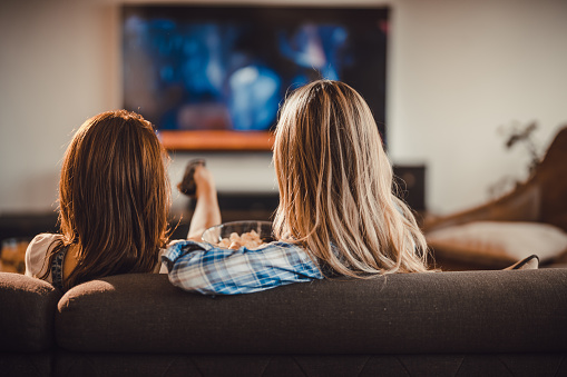 Back view of female friends watching TV on sofa at home.