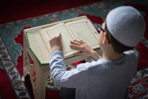 Rear View Of Muslim Kid Wear Skullcap Reading Quran On Prayer Mat At The Mosque