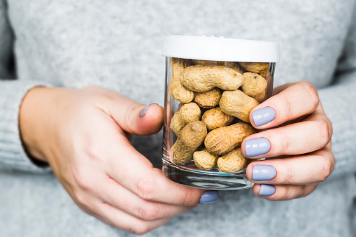 The woman is holding a jar with a white lid and peanut in the shell inside. Vegan and natural food concept