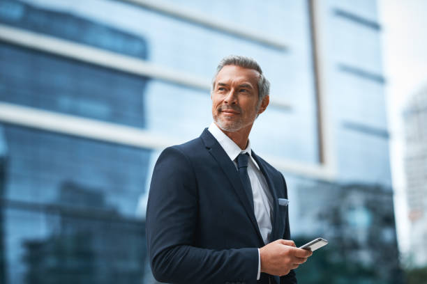 Hard work, determination, persistence creates a boss Shot of a handsome mature businessman in corporate attire using a cellphone outside outside during the day business man looking away stock pictures, royalty-free photos & images