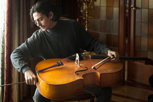 Young handsome white elegant long haired man with moustache preparing his cello to be played on an elegant environment. Musician rehearsal concept.