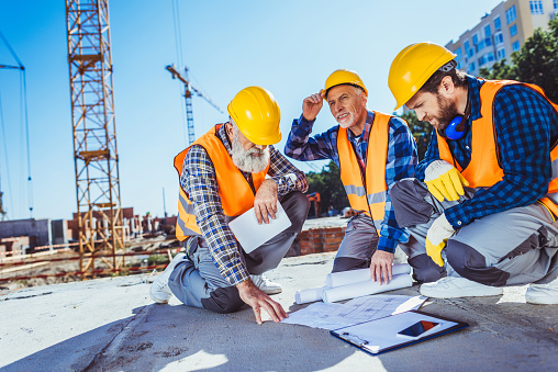 Three construction workers in uniform sitting on concrete at construction site, examining building plans