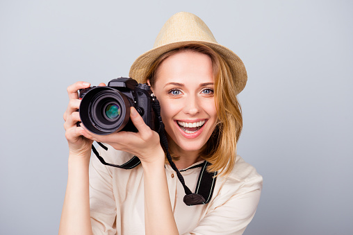 Young cheerful blond female photographer is smiling on the light blue background. She is excited and holding camera, wearing hat